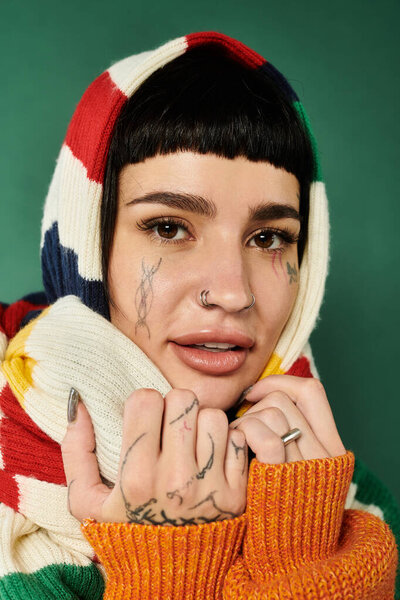 A young woman with vibrant tattoos smiles warmly while wearing a cozy sweater and scarf against a green backdrop.