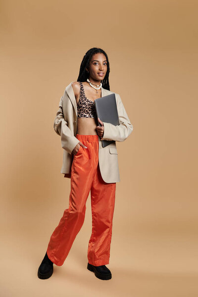 Молодая женщина уверенно стоит, украшенная в модную одежду, держа в руках ноутбук.