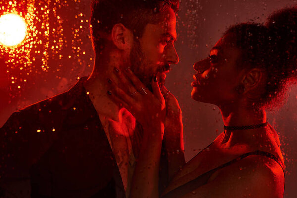 Пара разделяет интимный момент, освещенный мягким красным светом против капель дождя на стекле.
