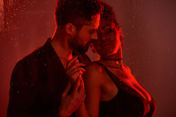 Пара разделяет нежное объятие, окруженное манящим красным свечением и нежным дождем на стекло..