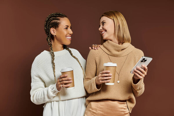 Две молодые женщины в осенних нарядах делятся радостными моментами с теплыми напитками в уютной обстановке.