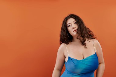 Göz kamaştırıcı makyajlı genç bir kadın parlak turuncu bir arka planda mavi elbisesiyle güzelliğini sergiliyor..