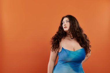 Genç bir kadın kendine güveni olan mavi bir elbise giyer, canlı turuncu bir arkaplan karşısında stilini ve makyajını sergiler..