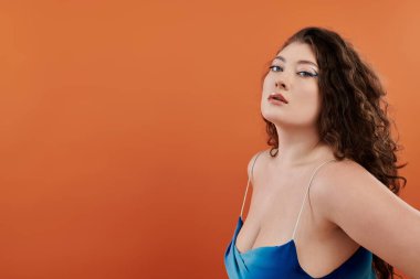 Kendinden emin genç ve iri yarı bir kadın canlı turuncu bir arkaplana karşı mavi bir elbise içinde zarifçe poz veriyor..