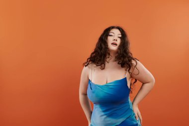 Genç, güzel, iri yarı bir kadın mavi bir elbiseyle turuncu bir arka planda kendine güvenerek duruyor..