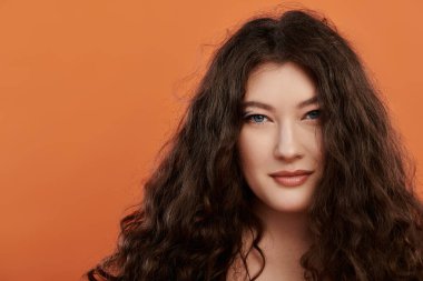 Göz kamaştırıcı genç bir kadın güzelliğini sergiliyor, turuncu arka planda kendine güveni yayıyor..