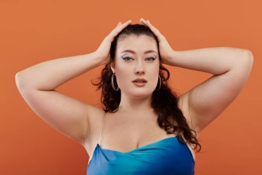 Çarpıcı mavi elbiseli kendine güvenen genç bir kadın turuncu arka planda poz veriyor ve güzelliğini sergiliyor..