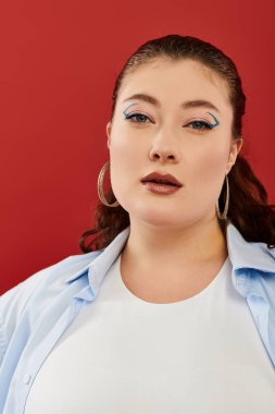 Göz kamaştırıcı, iri yarı bir kadın canlı makyajını, koyu kırmızı arka planda dururken sergiliyor..