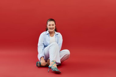 A joyful young woman showcases her bright makeup and style while sitting on a vivid red backdrop in a studio. clipart