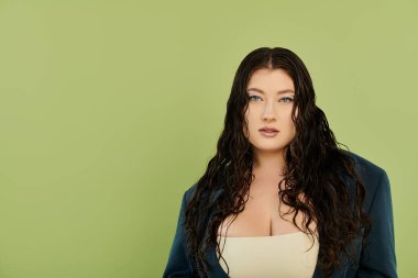 Güzel kıvırcık saçlı şık, iri yarı bir kadın yeşil arka planda özel dikim bir takım elbiseye olan güvenini sergiliyor..