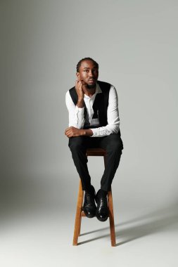 A young African American man sits thoughtfully on a stool, dressed in a sharp suit against a muted grey background. clipart