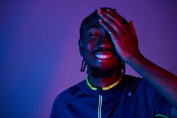 Молодой человек, одетый в модный наряд, демонстрирует сияющую улыбку, освещенную яркими неоновыми огнями.