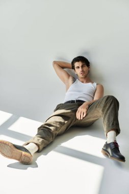 Dövmeleri olan şık bir genç adam gri bir zemine yaslanır. Kendine güveni ve cazibesi vardır..