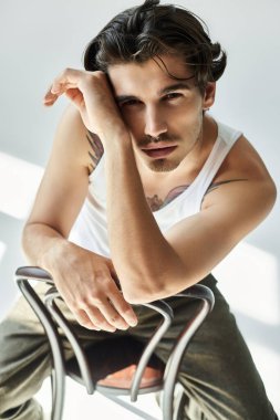 Askılı tişörtlü genç bir adam düşünceli bir pozla rahatlıyor, gri bir arka planda dövmesini ve çekiciliğini gösteriyor..