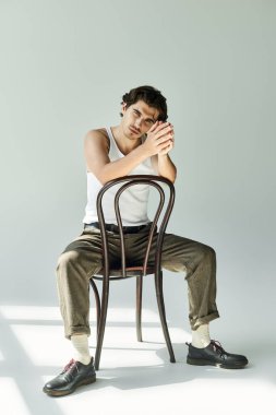Dövmeli genç bir adam, kolsuz tişörtle gevşiyor, düşünceli bir şekilde sandalyeye yaslanıyor ve sessiz bir arka plana yaslanıyor..