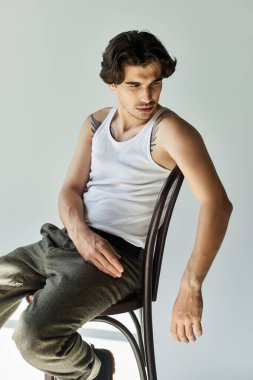 Kolsuz tişörtlü kendine güvenen genç bir adam sandalyede zarif bir şekilde otururken dövmelerini sergiliyor..