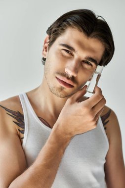 A young man with tattoos wears a tank top while thoughtfully gazing, framed by a simple grey background. clipart