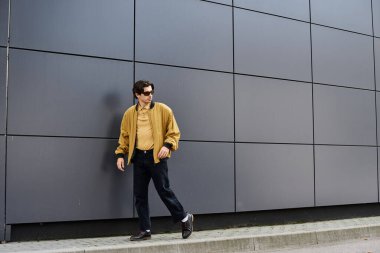 Modayı takip eden genç bir adam şık sonbahar kıyafetleriyle şık ve çağdaş bir duvarın önünde poz veriyor..