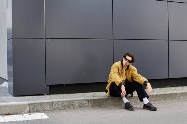 Genç bir adam, çağdaş mimarinin yanındaki kaldırımda güven içinde poz verirken sonbahar modasını sergiliyor..