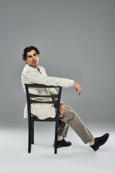 Молодой, красивый мужчина в элегантной формальной одежде сидит на стуле, излучая уверенность и очарование в студии.