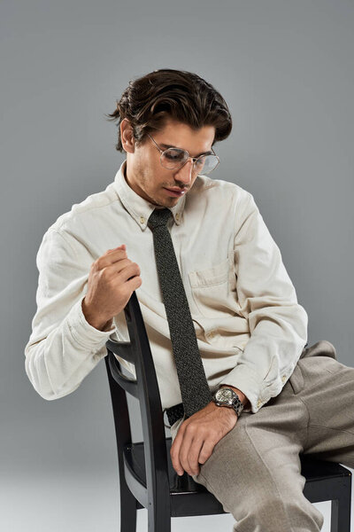 Молодой человек в модной формальной одежде занимает вдумчивую позу, сидя на сером фоне..