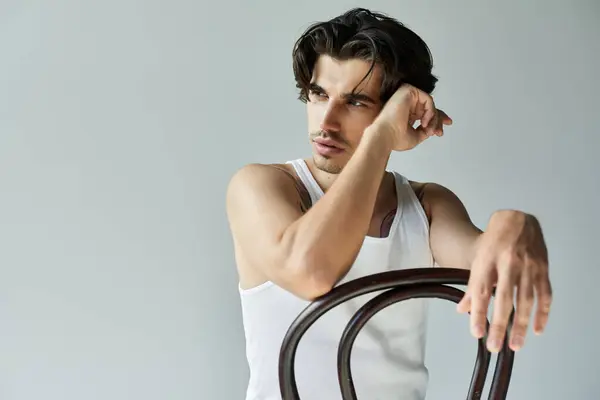 stock image A young man in a tank top leans against a chair, showcasing his tattoo while gazing thoughtfully into the distance.