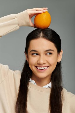 A cheerful young woman in trendy attire balances an orange atop her head, exuding joy. clipart