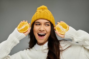 A joyful young woman sports a yellow beanie, holding fresh orange halves and smiling brightly. clipart