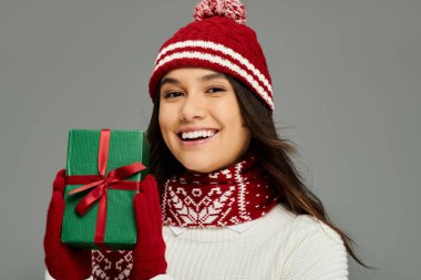 The woman beams with joy, showcasing a wrapped holiday gift in her cozy winter attire. clipart