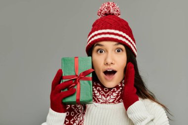 Excited woman holds a beautifully wrapped gift, expressing joy while dressed for winter festivities. clipart