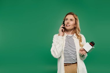 Güzel kıvırcık saçlı, sıradan giyinmiş bir kadın mikrofon tutarken telefonla konuşuyor..