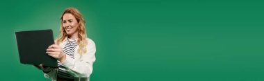Güzel bukleli ve sıradan gülüşlü bir kadın laptopunu canlı yeşil bir arka plana karşı kullanırken.