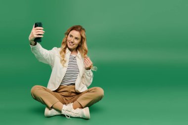 Sıradan giyinmiş neşeli bir kadın bağdaş kurup yeşil bir zeminde selfie anının tadını çıkarıyor..