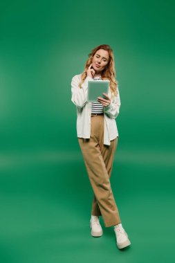 Kıvırcık saçlı şık bir kadın yeşil bir alanda tabletiyle uğraşırken düşünceli bir şekilde yansıtır..