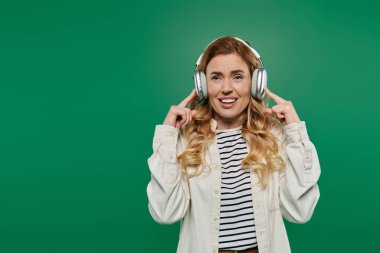 Kıvırcık saçlı mutlu bir kadın kulaklıklı müzik dinliyor, yeşil bir arka plana gülümsüyor..