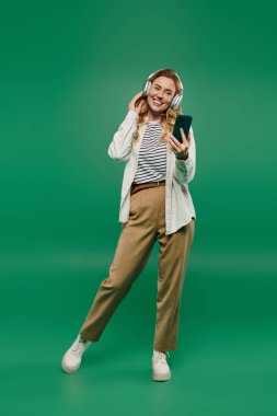 Kıvırcık saçlı neşeli bir kadın telefonunda müzik dinlerken gündelik kıyafetler giyiyor ve gülümsüyor..