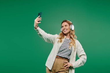 A joyful woman in casual wear takes a selfie with headphones, set against a bright green background. clipart