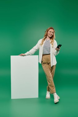 Gülümseyen bir kadın, elinde akıllı bir telefonla canlı yeşil bir arka plana karşı durup neşe saçar..