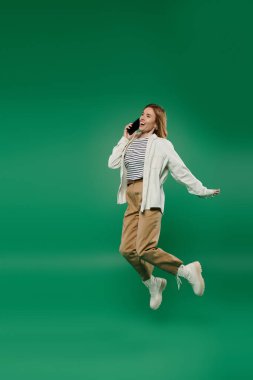 Günlük kıyafetli neşeli bir kadın telefonuyla konuşurken neşeli bir şekilde zıplıyor, pozitif enerji yayıyor..