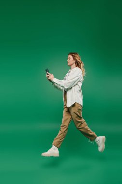 Gündelik giysiler içinde güzel bir kadın, canlı yeşil bir zemin üzerinde, akıllı telefonuna odaklanmış bir şekilde ileri doğru yürüyor..