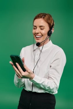 Resmi giyinmiş neşeli bir kadın, kulaklık ve akıllı telefon kullanarak müşterileriyle neşe içinde iletişim kurar..
