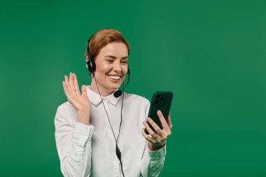 Resmi bir kadın, parlak yeşil bir arka planda telefonuyla iletişim kurarken gülümser ve el sallar..