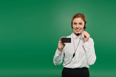 A confident woman in a white shirt smiles while holding a device, ready for call center communication. clipart