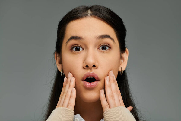 An emotional young woman touches her face with surprise, showcasing her stylish outfit.