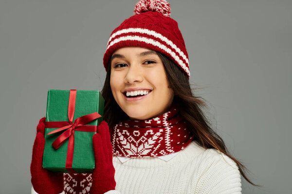 The woman beams with joy, showcasing a wrapped holiday gift in her cozy winter attire.