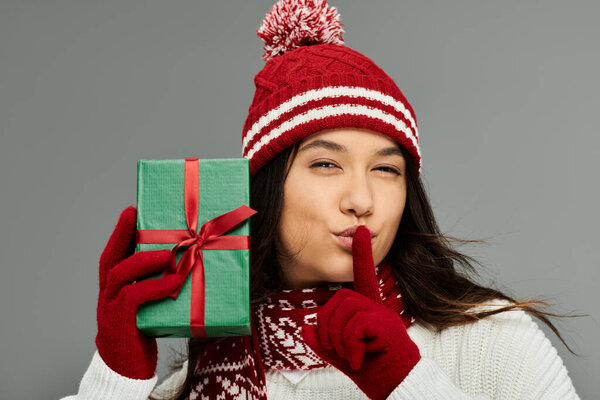 Молодая женщина в стильном зимнем наряде игриво держит подарок, сигнализируя о тишине с улыбкой.