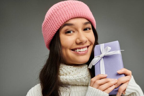A young woman wearing a pink beanie and scarf beams with happiness while holding a present.