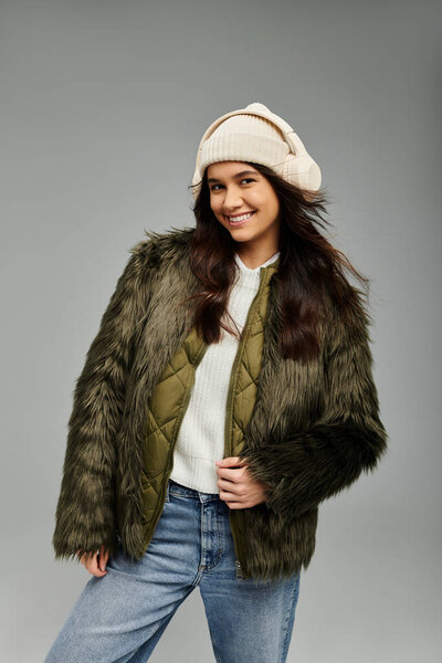 Молодая женщина в стильной зимней одежде тепло улыбается, позируя на сером фоне..