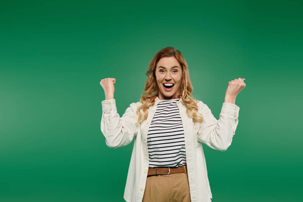 Веселая женщина с вьющимися волосами выражает волнение во время ношения повседневной одежды в ярко-зеленой обстановке.