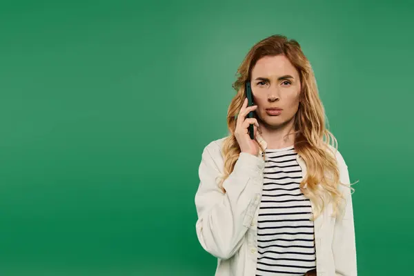 stock image A woman with curly hair in casual attire appears deep in thought while talking on her phone amid a green backdrop.